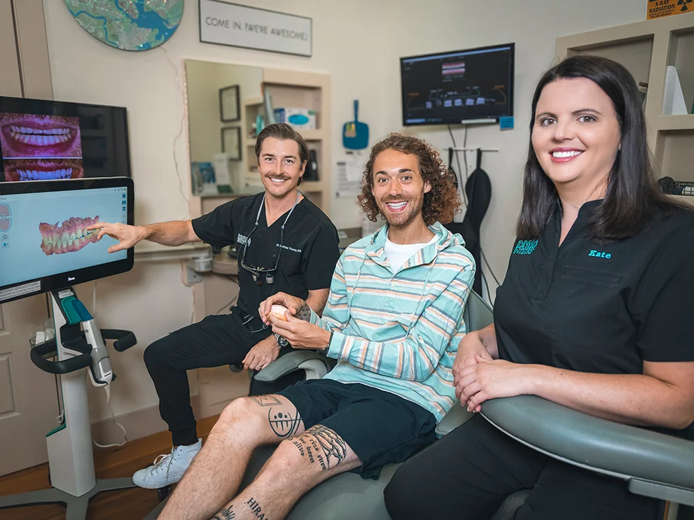 Dentist pointing at a 3D dental scan on a monitor while seated beside a smiling patient holding a dental model, with a dental assistant also smiling and seated nearby in a modern dental operatory. The screen displays digital renderings of teeth and gums.
