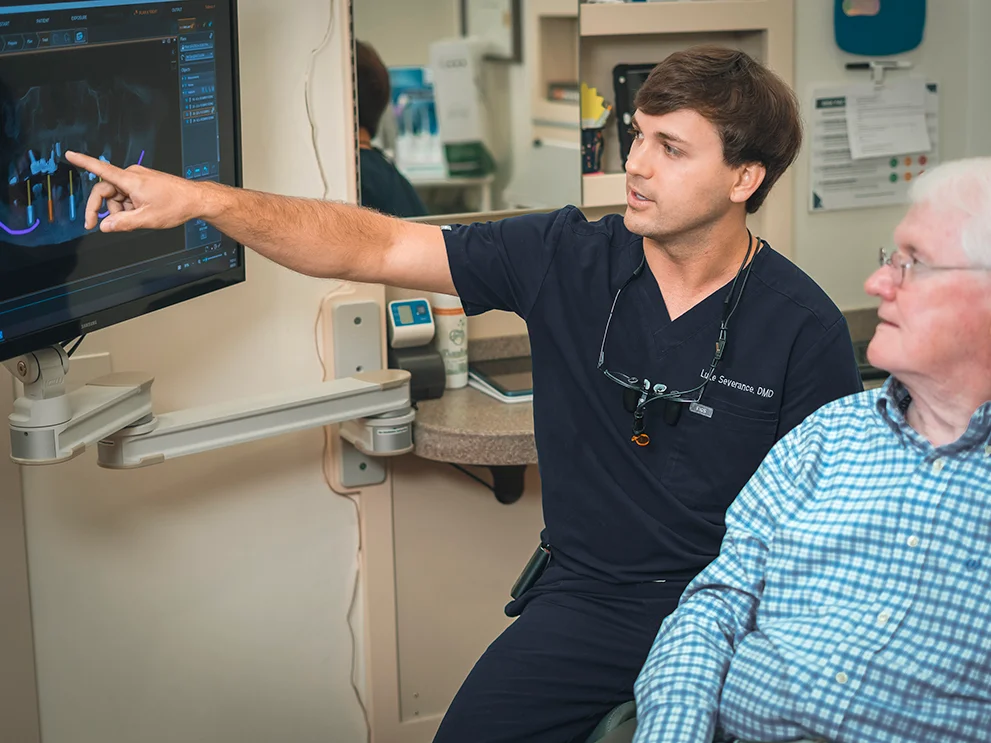 Male dentist in navy scrubs pointing at a dental X-ray on a monitor while explaining treatment to an older male patient seated beside him in a dental operatory. The screen displays an image with dental implants and jaw structures.
