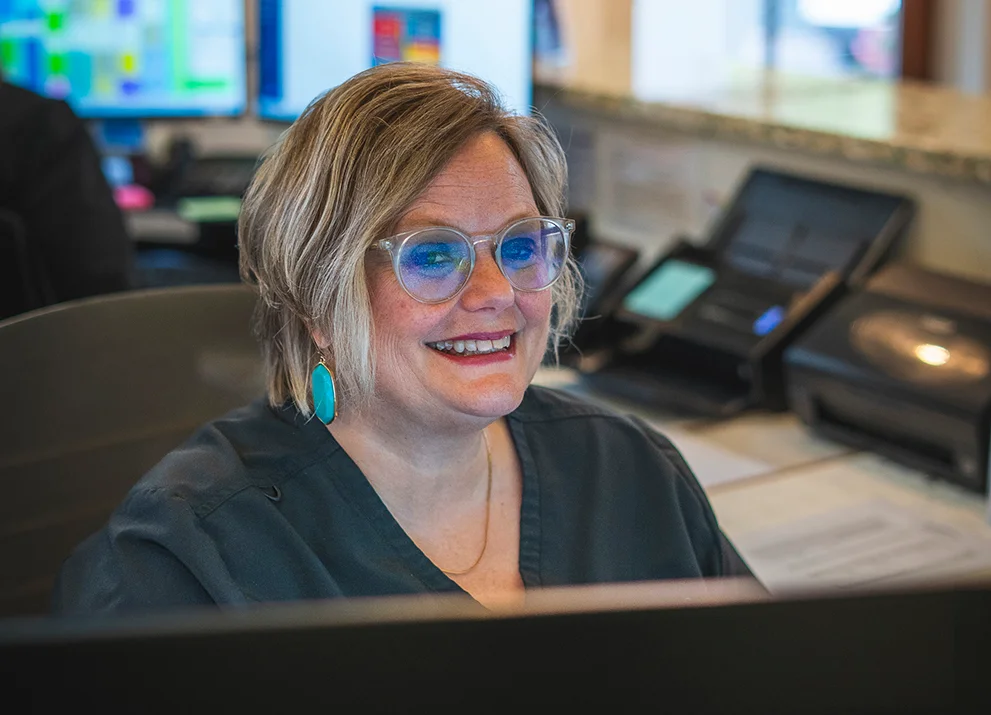 Smiling dental office receptionist wearing glasses and turquoise earrings, seated at a front desk with multiple monitors and office phones in the background.